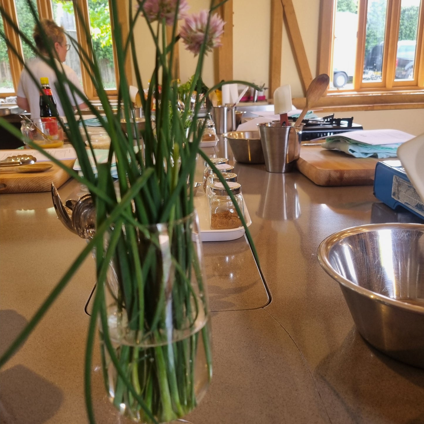 The Greek Kitchen by Event sits on a kitchen counter with bowls, utensils, and ingredients—perfect for preparing authentic Greek food as sunlight streams in through large windows.