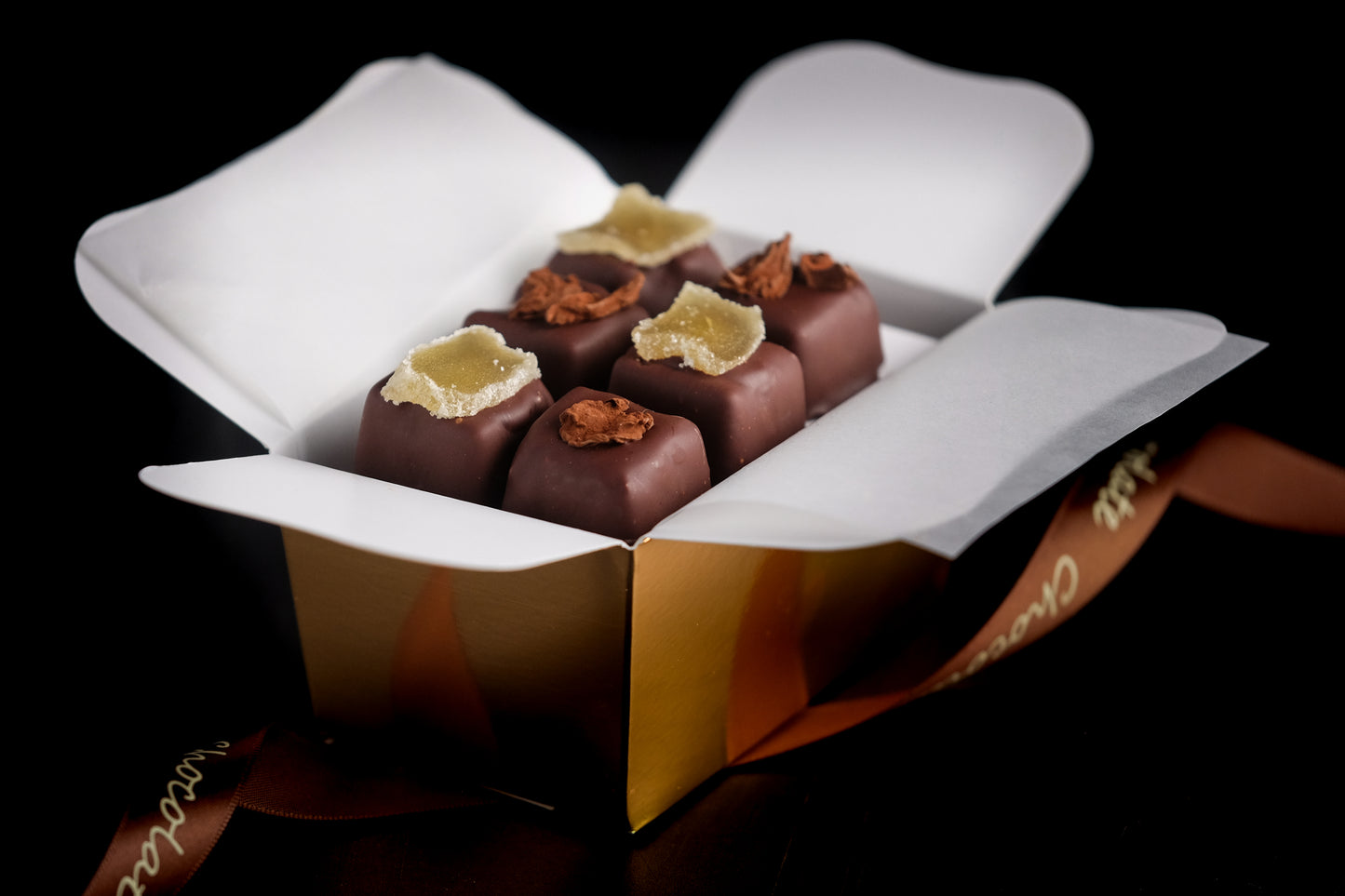 The Event Chocolate Experience features six square truffles in a gold box lined with white paper, topped with candied ginger and chocolate shavings. A ribbon accent completes this elegant presentation.