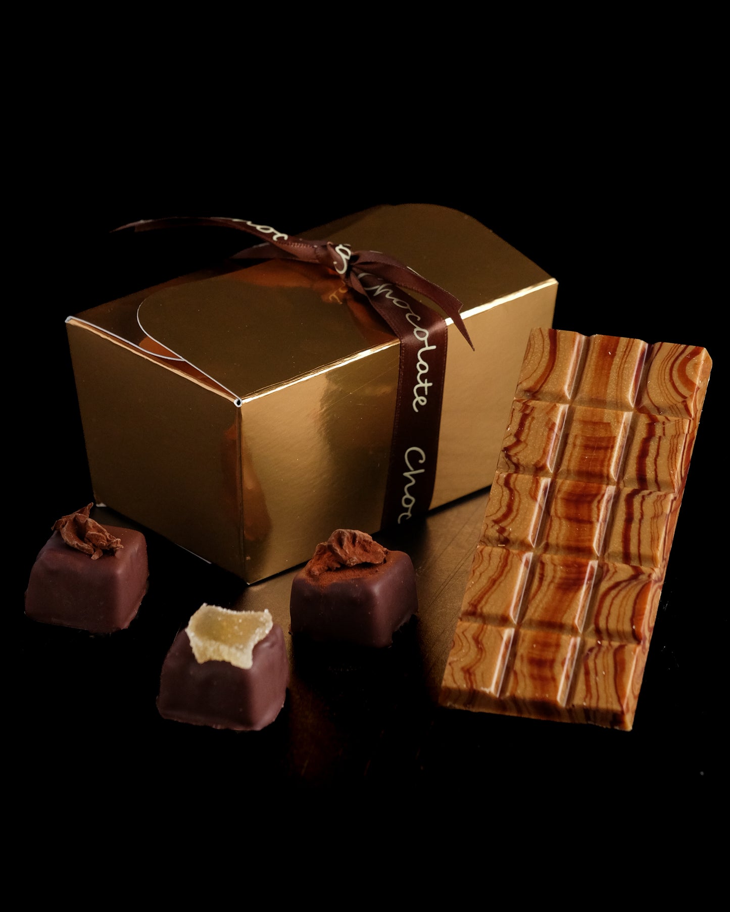 The Chocolate Experience by Event features a gold gift box with a brown ribbon, a marbled chocolate bar, and three assorted truffles—all beautifully displayed on a sleek black surface.