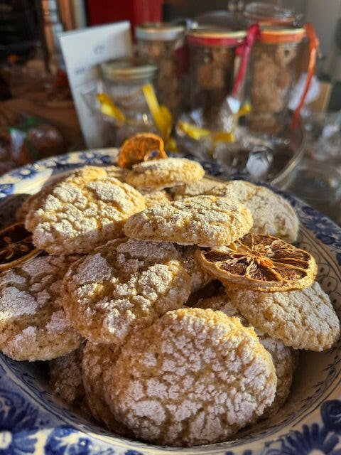 A decorative bowl from Event’s Edible Christmas Gifts...Buon Natale, as they say in Italy holds powdered sugar-dusted Lemon Ricciarelli cookies and dried citrus slices, with jars of holiday ingredients blurred in the background.