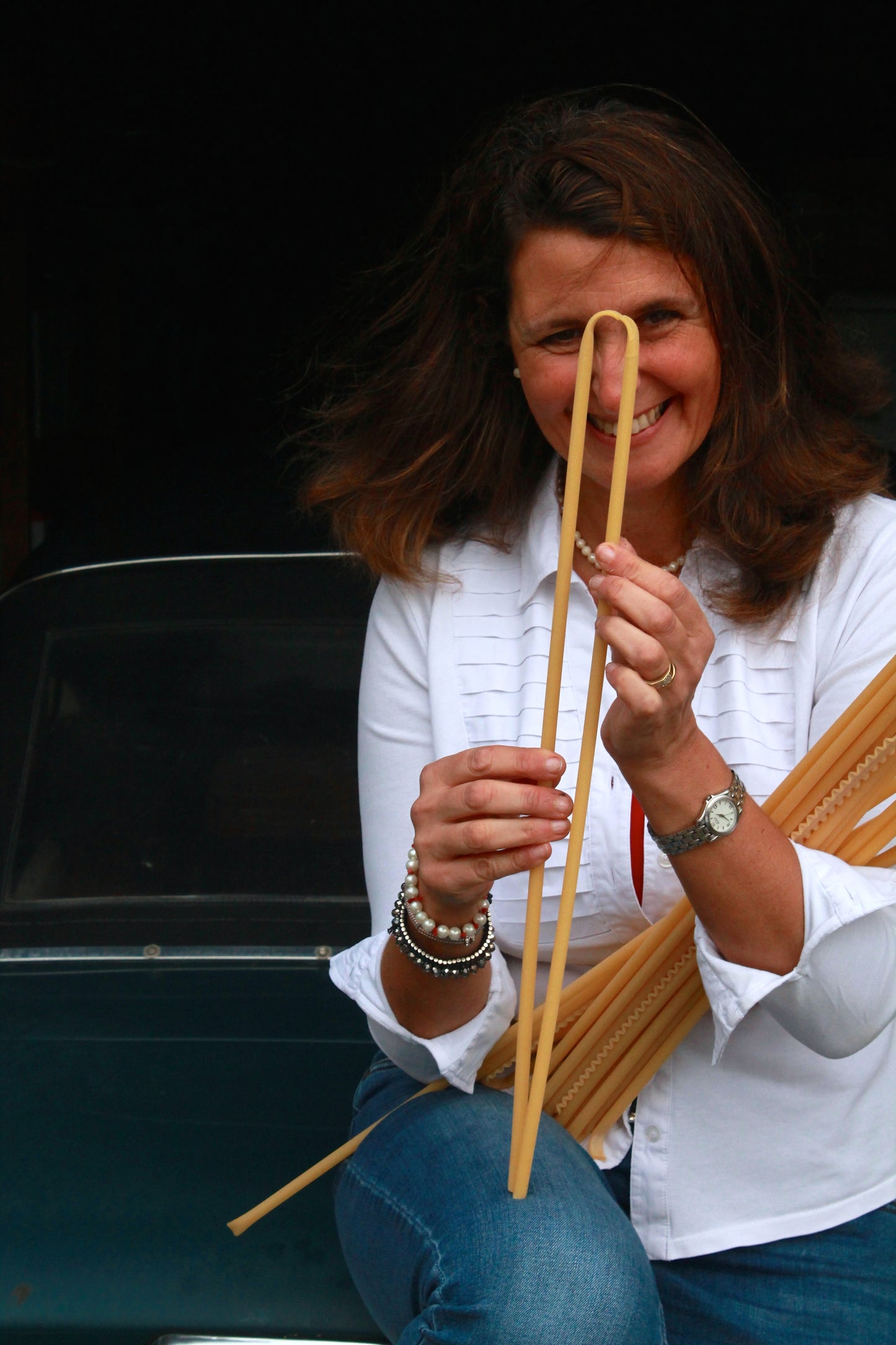 A woman in a white shirt and jeans smiles while holding long uncooked pasta, one piece arched—perhaps planning to enjoy it with Event’s Panettone Brioche e Crema di Limoncello for a delightful meal.