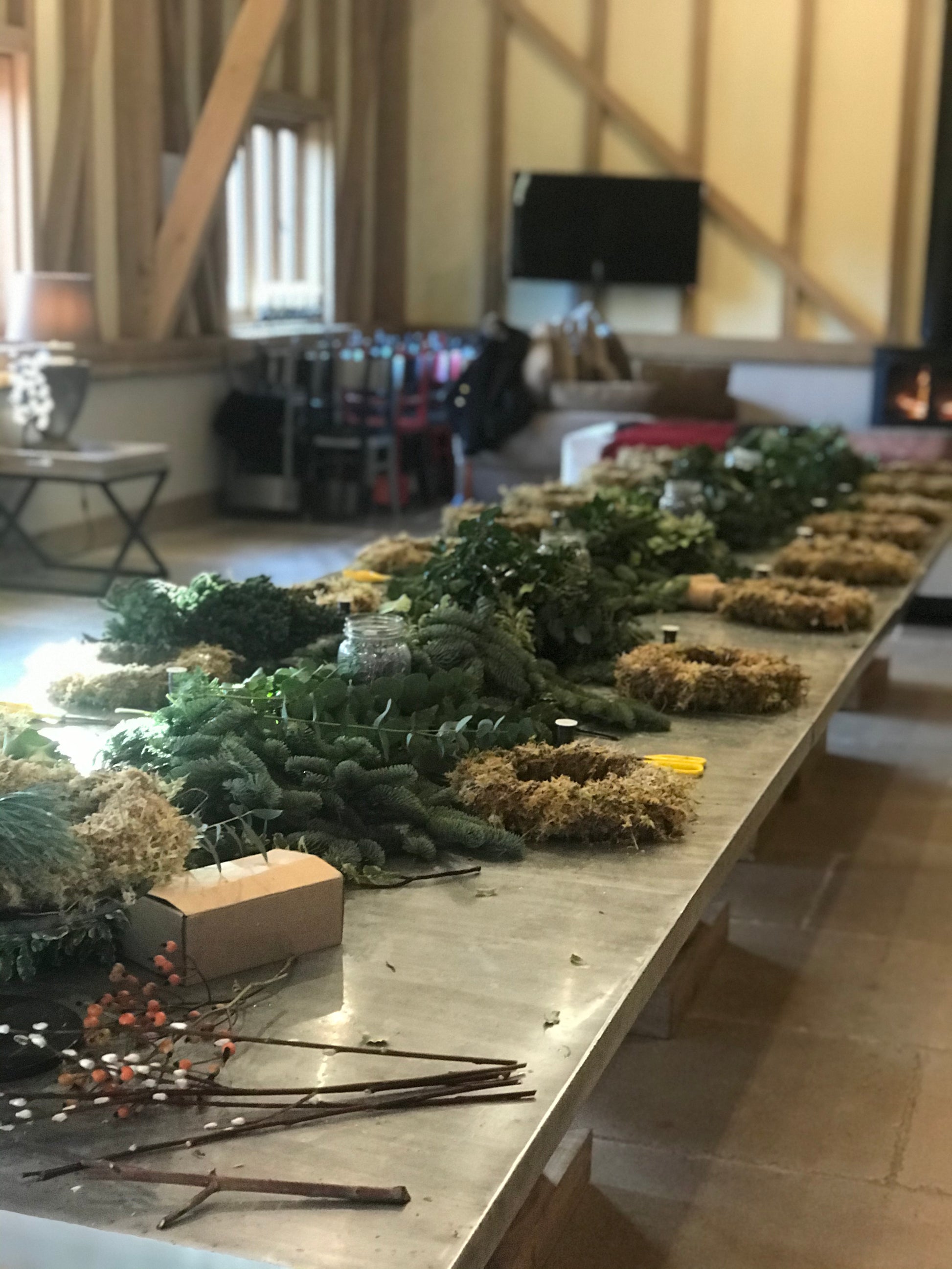 The Christmas Wreath Workshop by Event features a long table filled with greenery, moss, and craft supplies in a warmly lit room with wooden beams, stacked chairs, and a glowing fireplace in the corner.