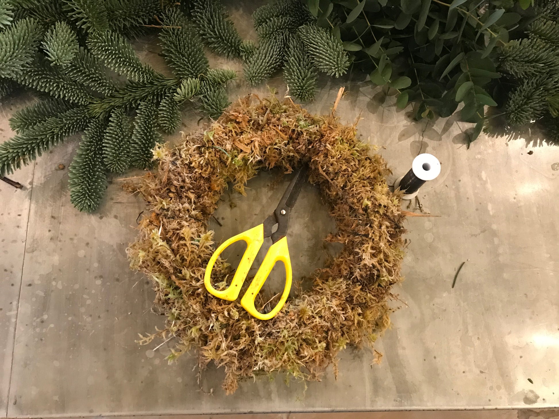 A yellow-handled pair of scissors, dried moss wreath, and black thread spool from Events Christmas Wreath Workshop are arranged with greenery on a wooden surface—ideal supplies for festive crafting.