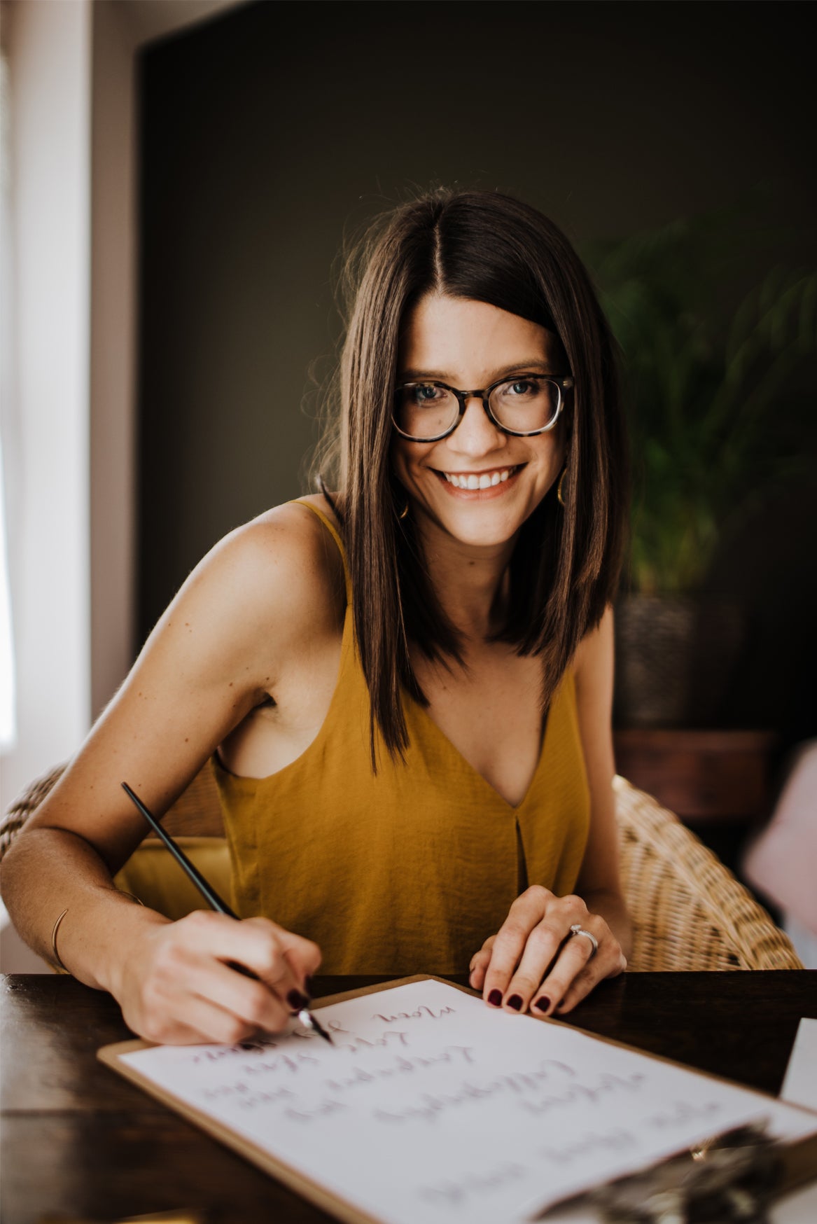 A woman with long brown hair and glasses, in a mustard yellow sleeveless top, smiles while writing on a clipboard at a wooden table—perhaps planning her next beginner project with Modern Calligraphy by Event. A potted plant sits in the background.