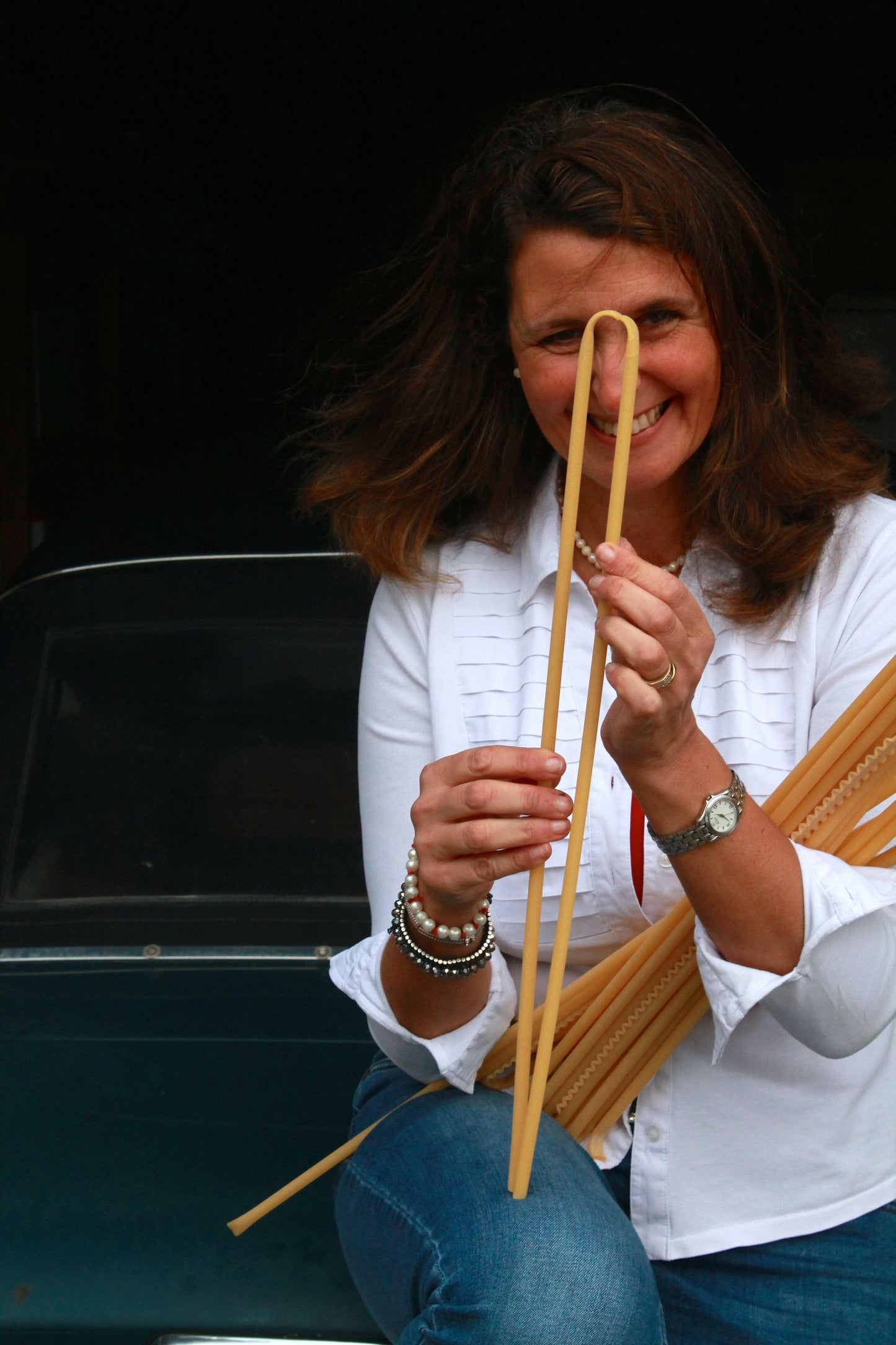 A woman in a white shirt and jeans sits before a car, smiling as she holds uncooked pasta, her joy capturing the spirit of Event's "The Amalfi Coast" experience—an ode to authentic Italian cooking in the sun.