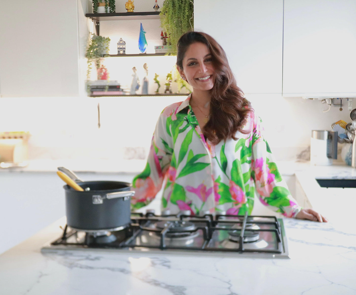 A woman in a floral shirt smiles in a modern white kitchen behind a stove with a pot, ready to teach Punjabi Comforts, Flavours of Home by Event. Plants and decor appear on open shelves in the background.