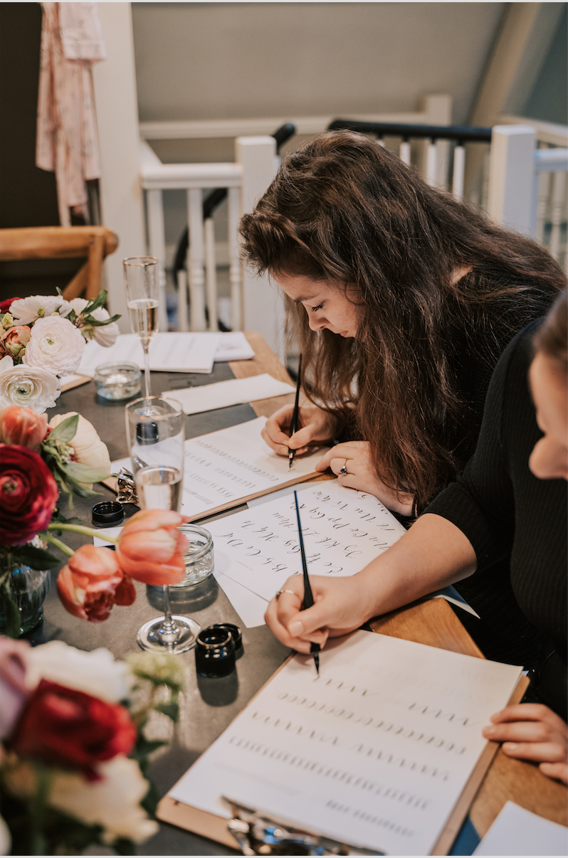 Two women enjoy practicing beginner Modern Calligraphy by Event at a cozy table, using brush pens, paper, ink pots, and surrounded by flowers and decorative stationery. Champagne glasses add to the inviting indoor atmosphere.