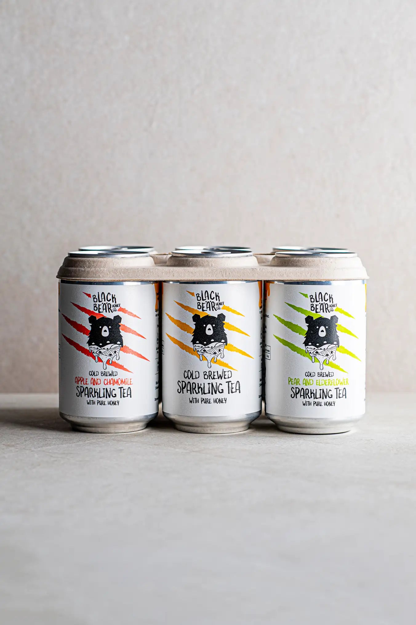 Three cans of Honesty Cold Brewed Sparkling Tea with Pure Honey—apple & chamomile, classic, and elderberry—are side by side on a neutral background. Each can displays a black bear illustration with vibrant claw marks.