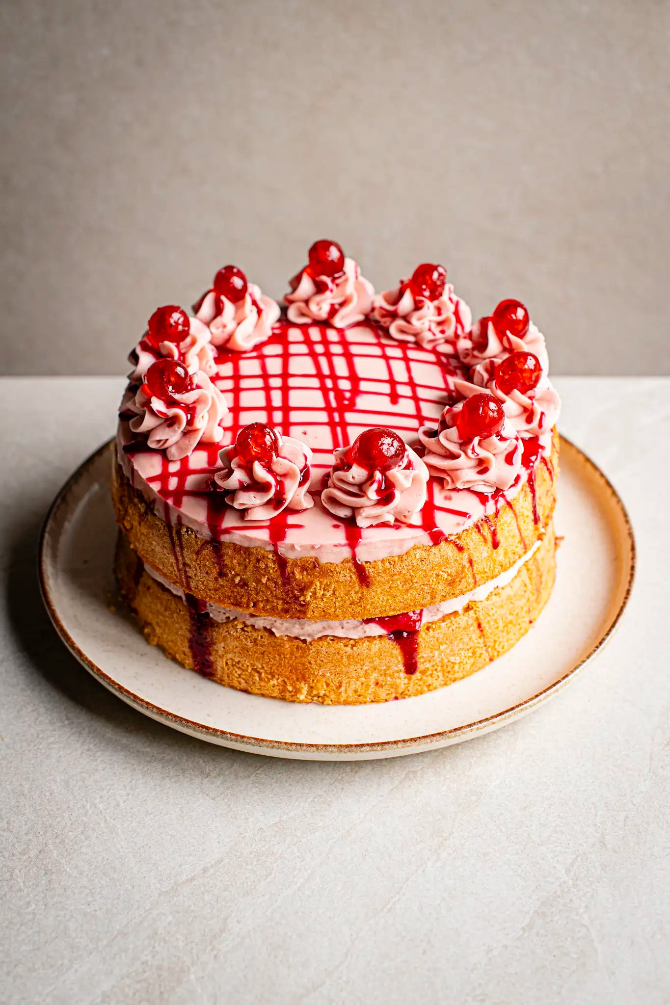 The Honesty Cherry Cake, a handmade two-layer cake with pink frosting, cream swirls, red cherries, and red syrup drizzles, is served on a white plate against a neutral background.