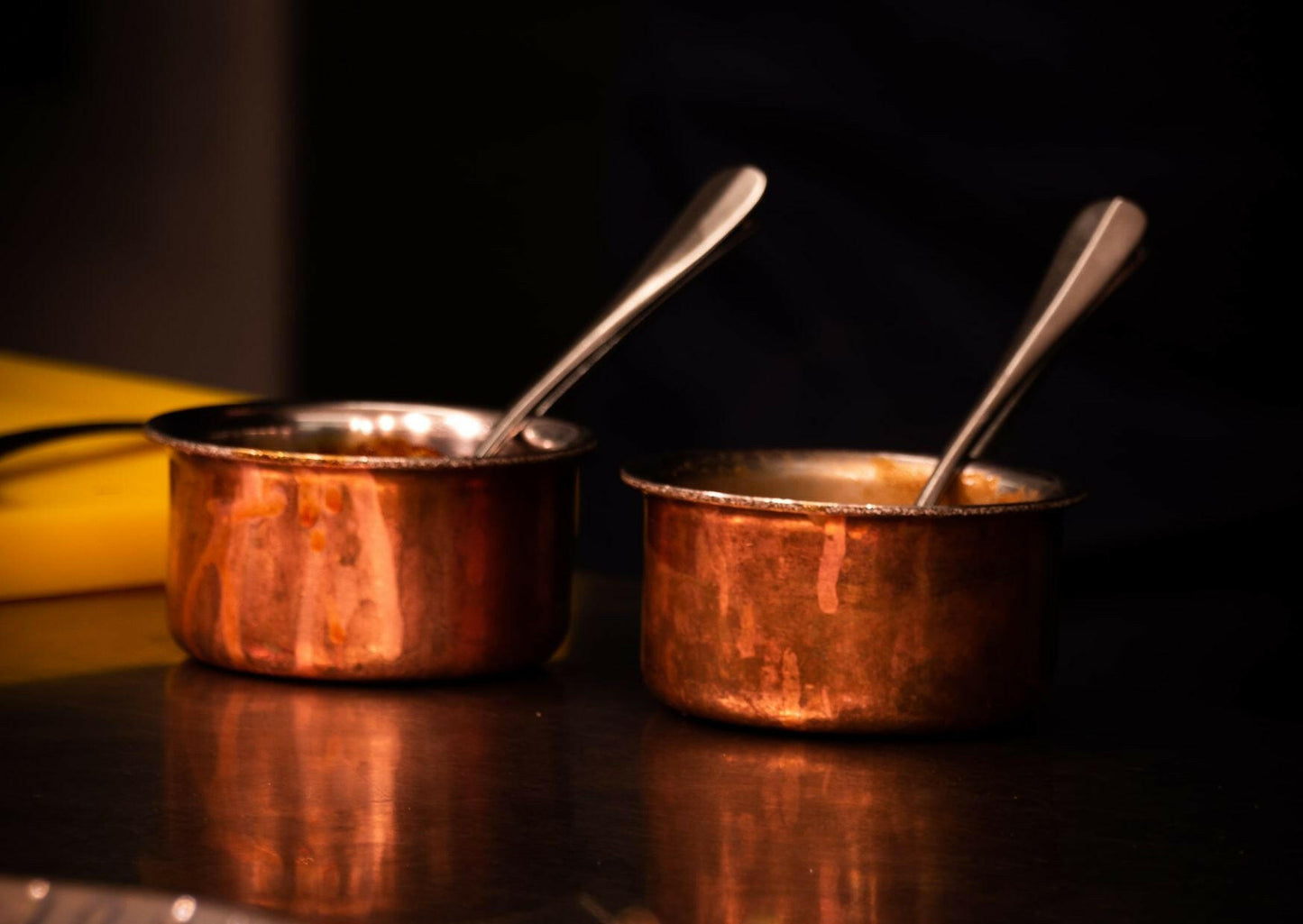 Two small copper pots from Event's "The Amalfi Coast" sit side by side on a reflective surface, each with a metal spoon inside, evoking authentic Italian cooking. The dark, blurred background adds an elegant touch.