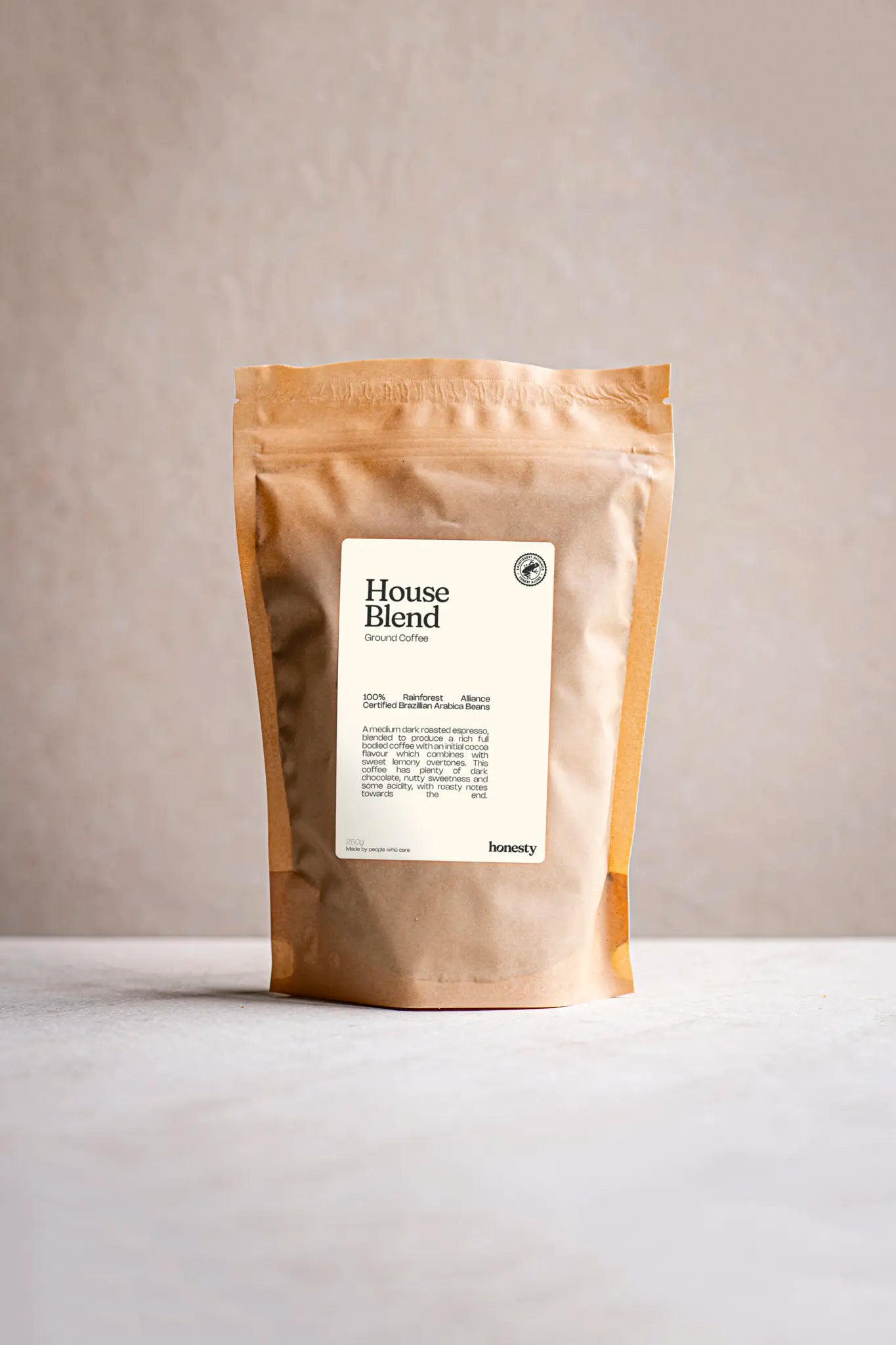 A brown, resealable coffee bag labeled House blend ground coffee by Honesty stands upright on a light surface against a neutral beige background. The minimalist label in black font highlights this handmade selection.