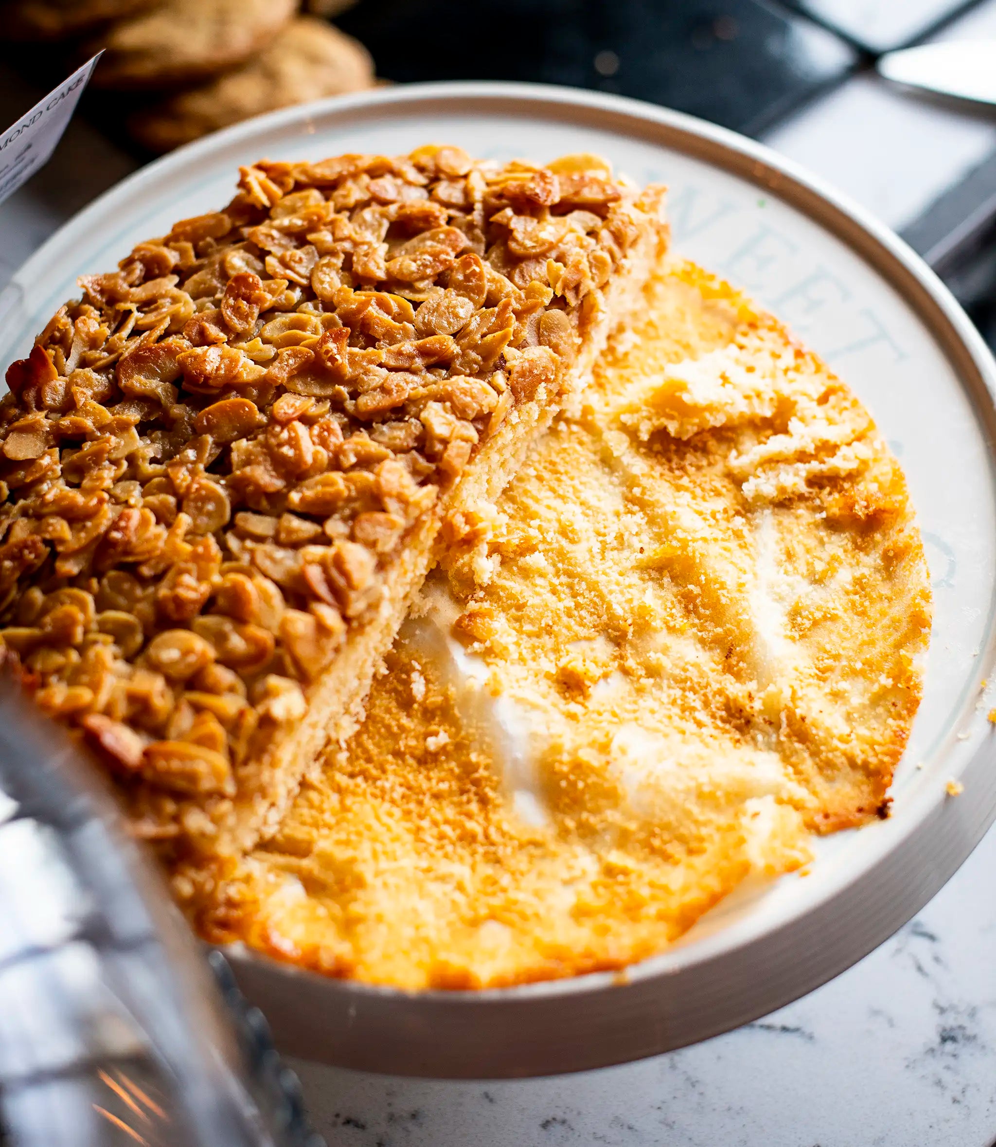 A round cake with a caramelized oat and nut topping sits on a white plate. Almost half of the cake has been eaten, revealing a moist, yellow interior. Crumbs and a few melted spots remain on the plate.