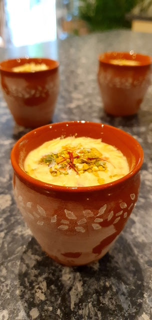 Three Flavours of India clay cups by Event, filled with creamy Indian dessert and topped with nuts and saffron, sit on a mottled gray countertop. The focused front cup highlights the rich, home-style inspiration behind this indulgent treat.