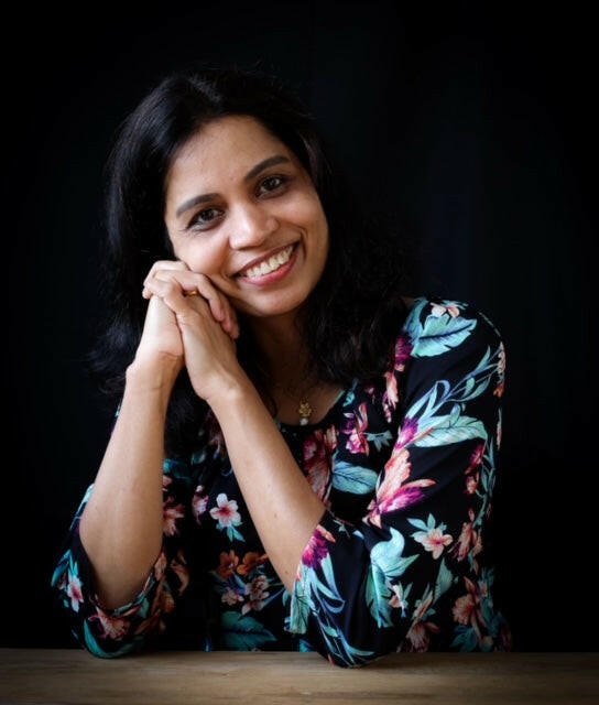 A woman with long dark hair, wearing a floral-patterned top, smiles warmly at a table against a dark background as she enjoys the Pancake Day Special - Dosa Masterclass by Event, savoring classic South Indian flavors.
