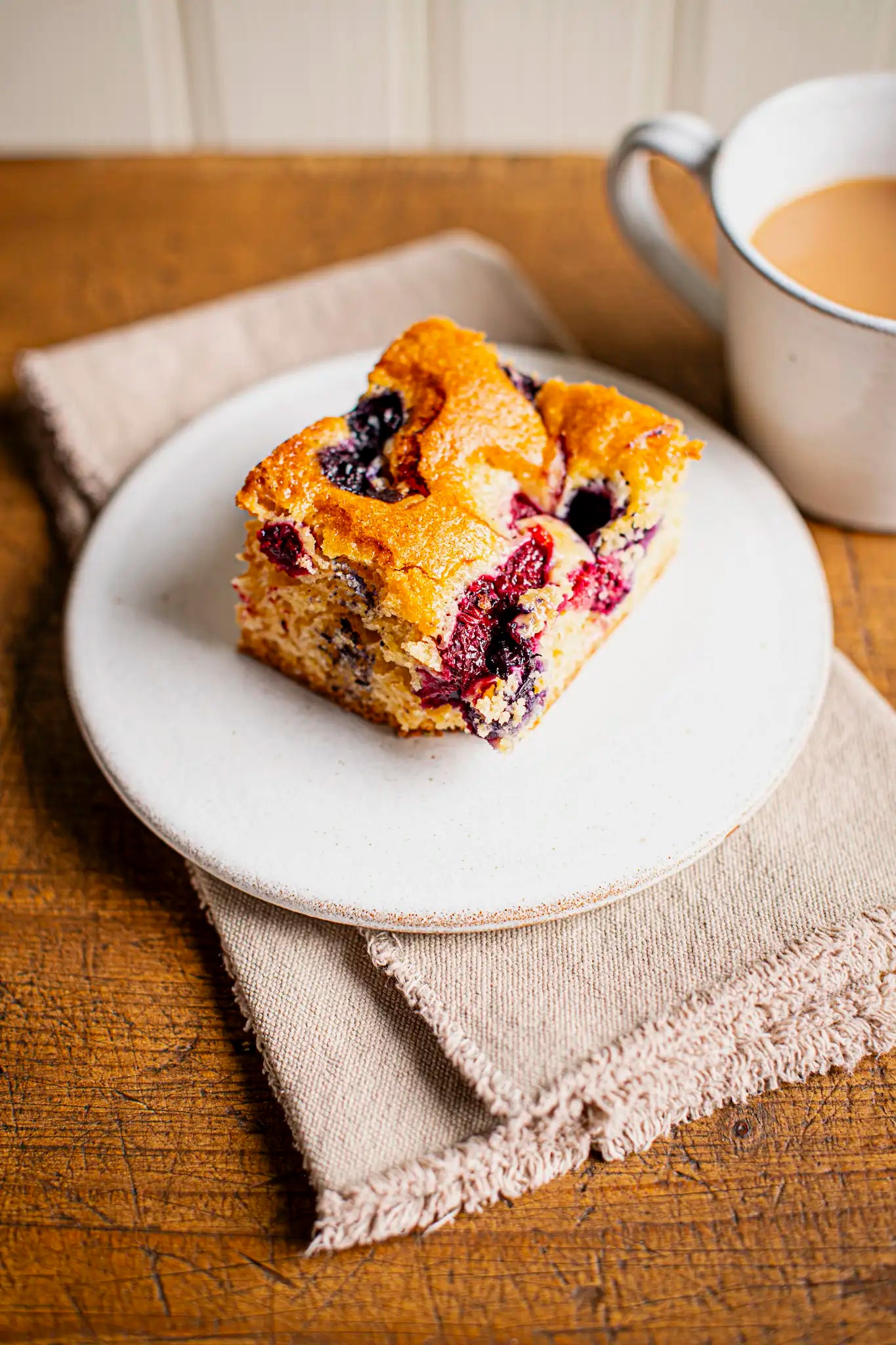A square piece of berry coffee cake sits on a white plate, placed on beige napkins atop a wooden table. A white mug filled with coffee or tea is in the background.