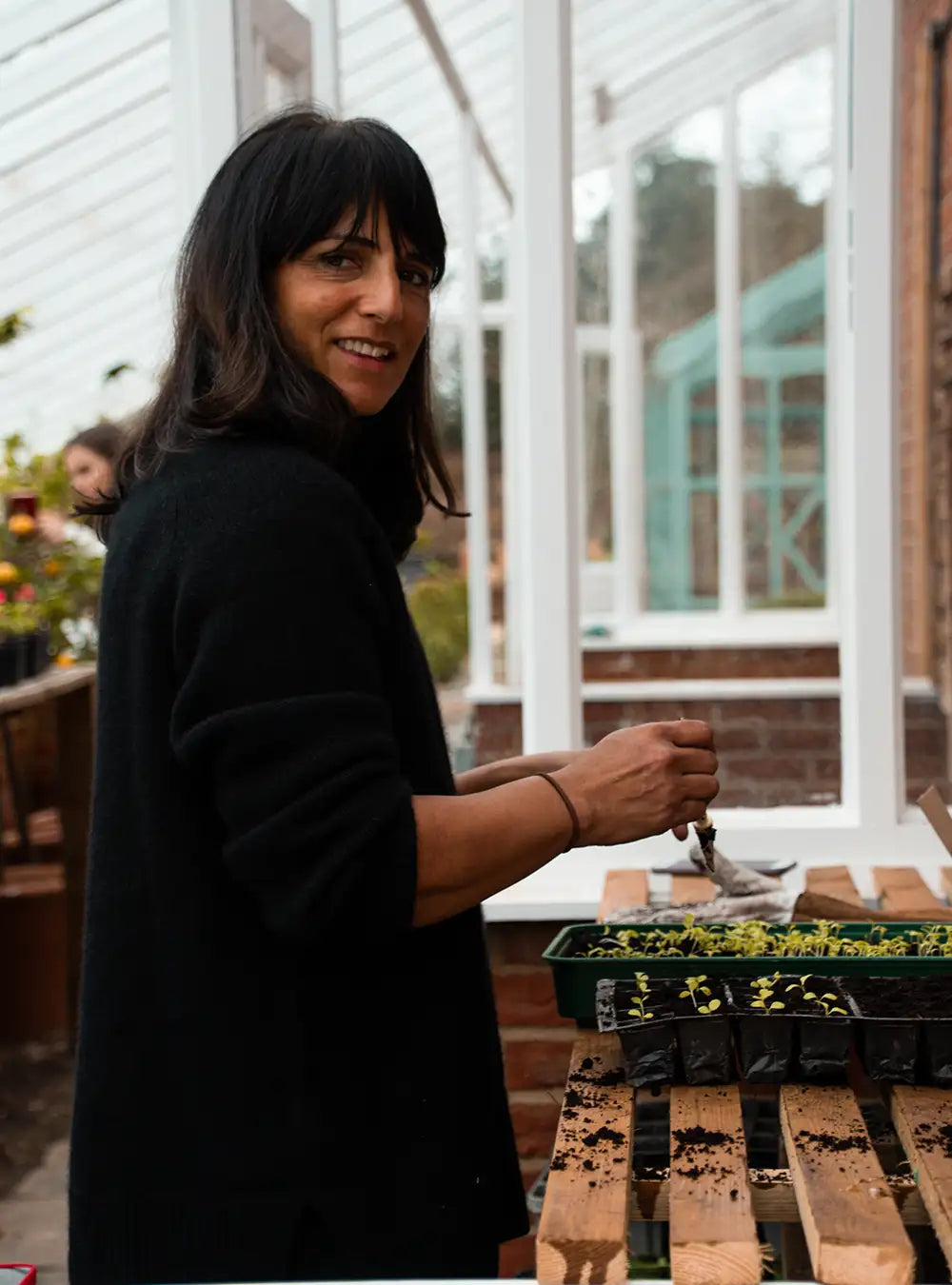 A woman with dark hair and a black sweater smiles while tending to young plants in trays inside a bright greenhouse.