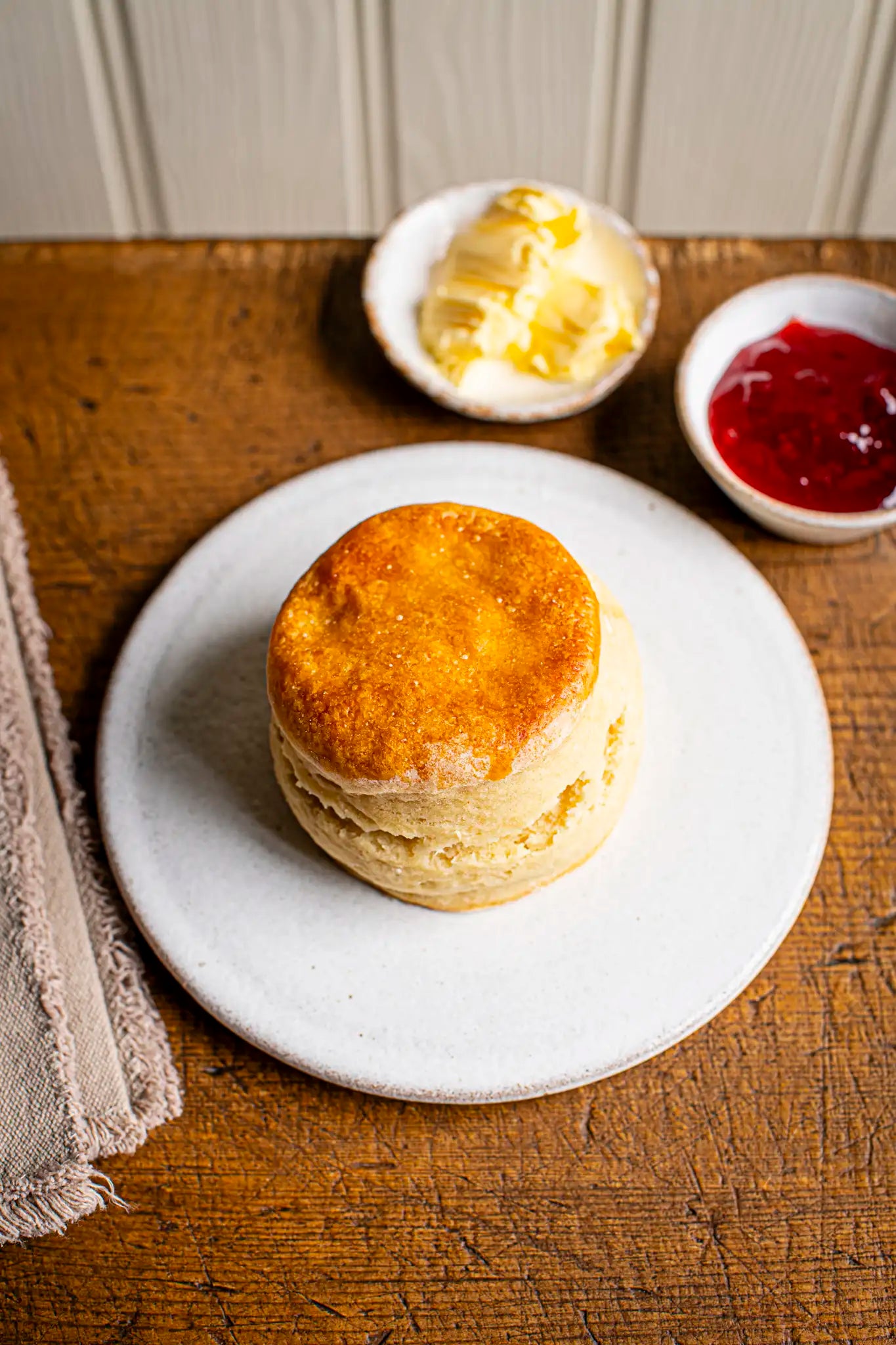 A golden sponge cake, Honesty’s Victoria Sponge, rests on a white plate with small dishes of butter and red jam in the background on a wooden table.