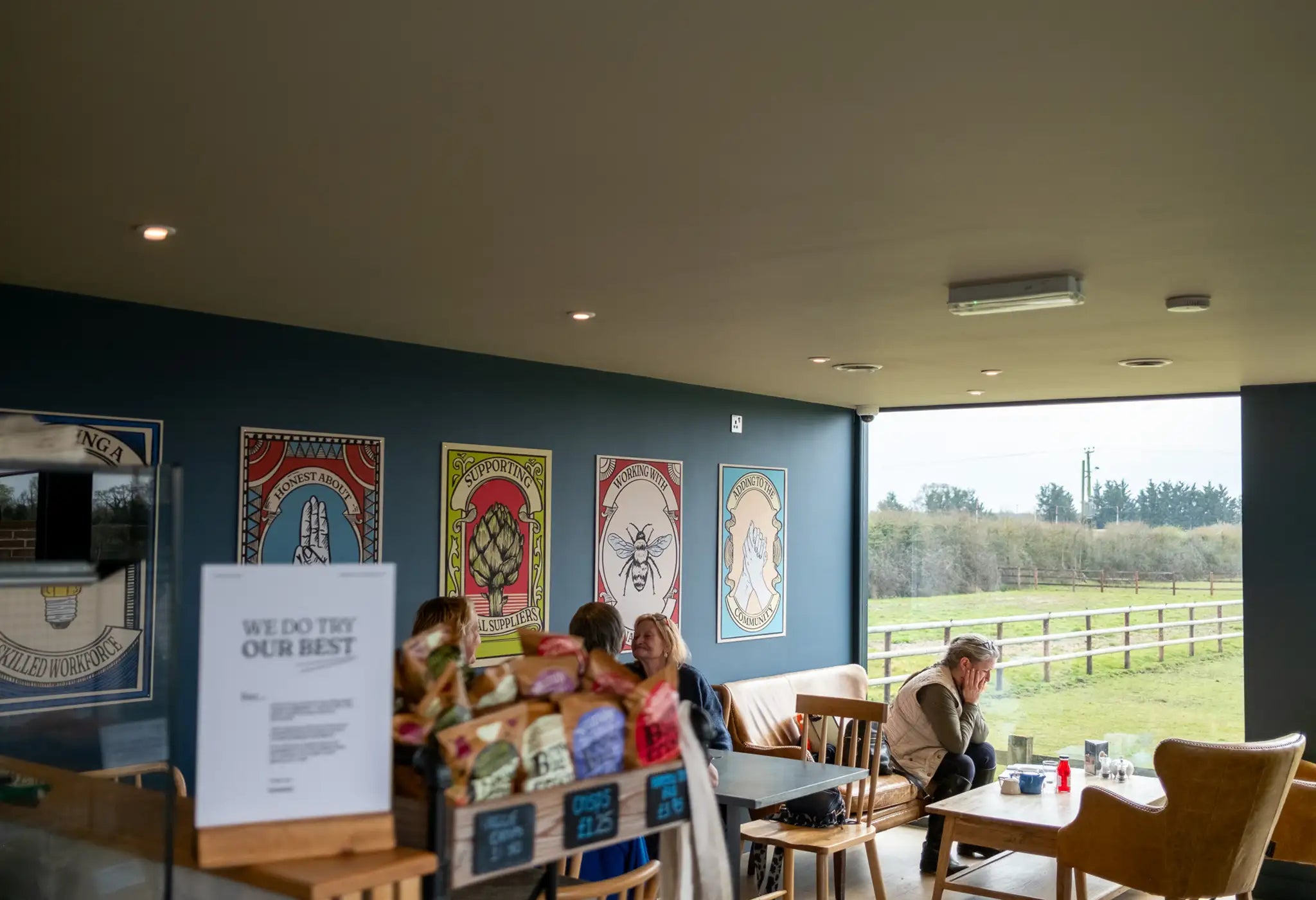 People sit at tables inside a cozy cafe with large windows overlooking a grassy outdoor area. Colorful artwork decorates the dark blue wall, and a display of snacks sits in the foreground.