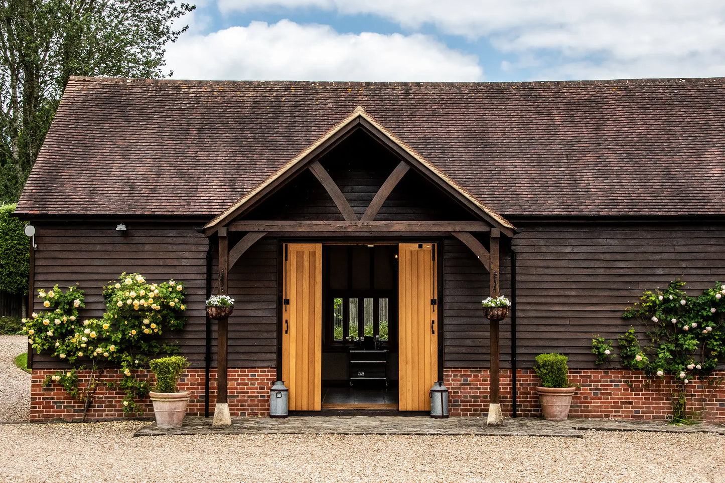 A rustic wooden barn with a brick base, open double wooden doors, and hanging flower baskets, surrounded by potted plants on a gravel courtyard under a partly cloudy sky.
