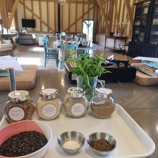 A kitchen counter for Event’s The Magic of Sri Lankan Cuisine features jars of coriander seeds, sesame seeds, ground white pepper, a small mint bouquet, metal bowls, and a tray. In the background: a spacious sunlit living area with wooden beams.