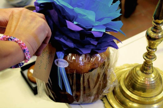 A hand holds a gift tag attached to Events Panettone Brioche e Crema di Limoncello, wrapped with blue and purple tissue paper, placed next to a brass lamp on a white surface.
