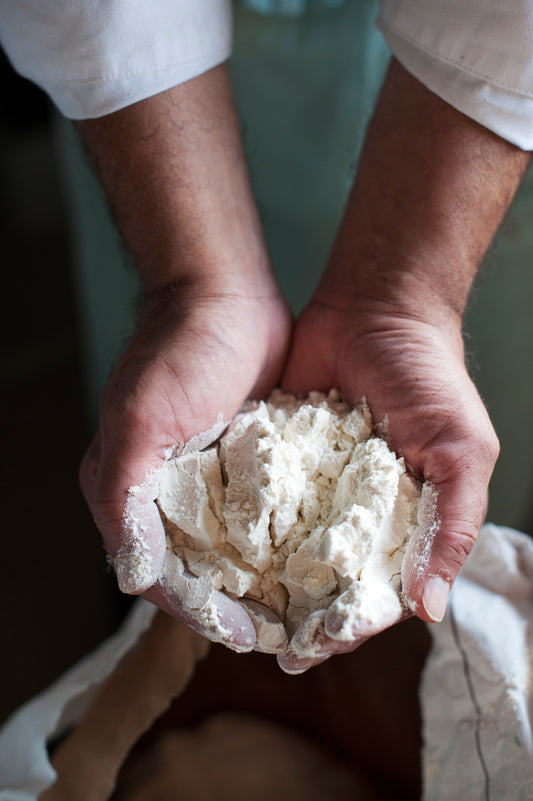 A close-up of two hands holding loose, white flour above an open sack, ideal for baking with Beautiful Breads by Event. The person wears a white long-sleeve shirt.