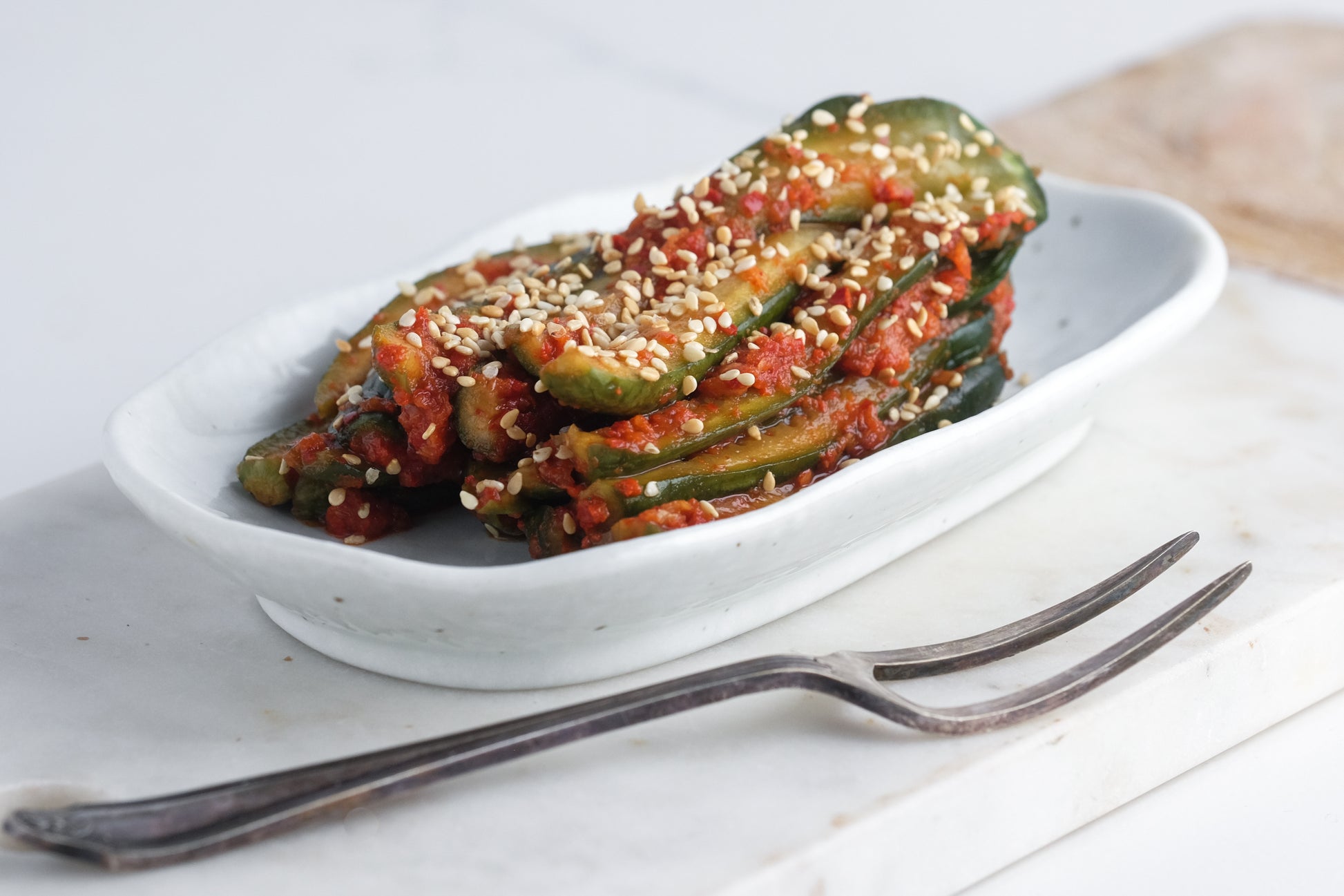 A small white dish of sliced cucumbers in red chili paste and sesame seeds showcases the vibrant flavors explored at the Event Fermented Foods Workshop. A metal fork sits nearby on a white surface, ready to enjoy.