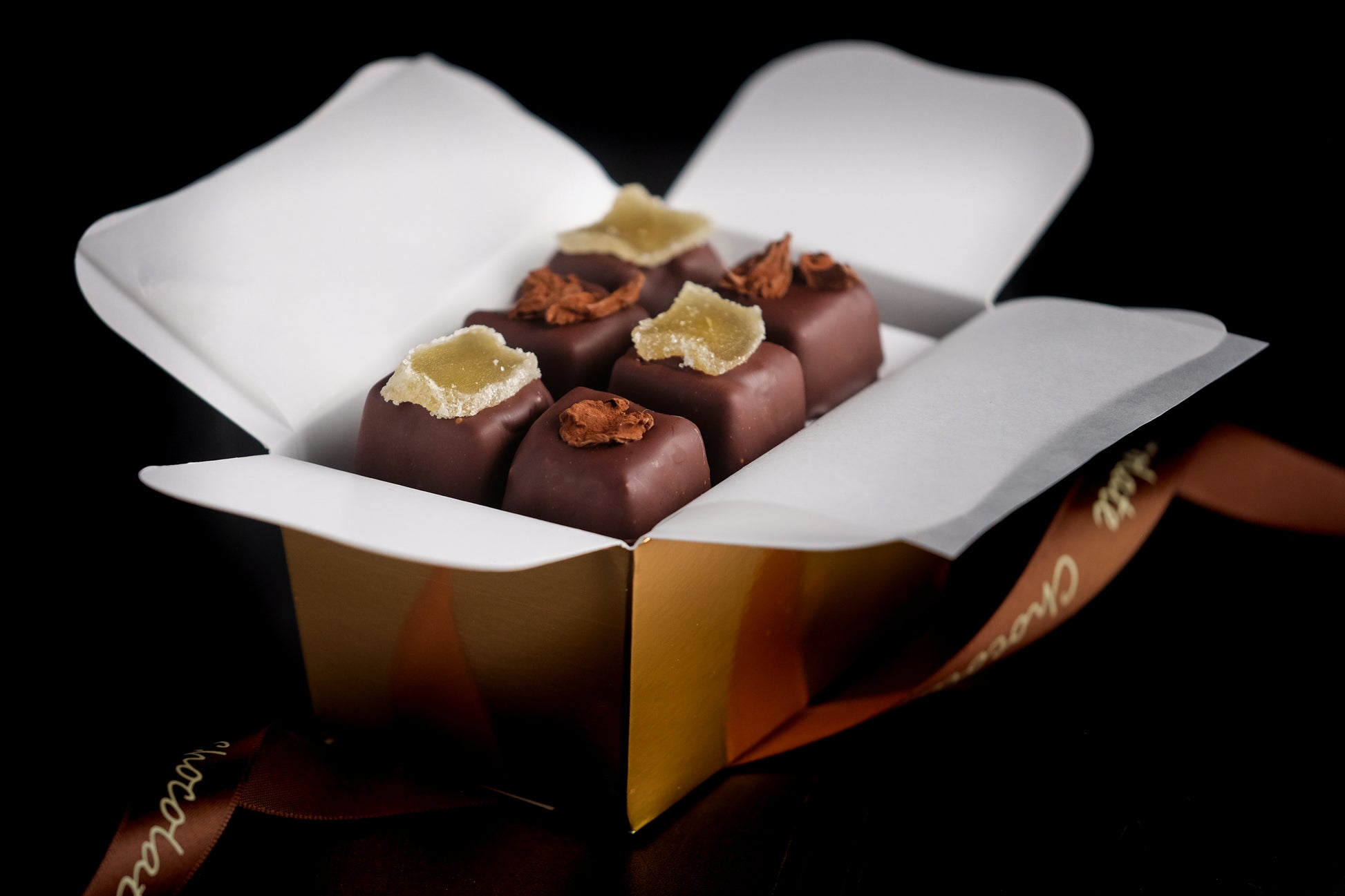 The Event Chocolate Experience features six square truffles in a gold box lined with white paper, topped with candied ginger and chocolate shavings. A ribbon accent completes this elegant presentation.