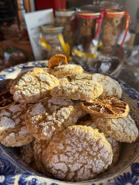 A decorative bowl from Event’s Edible Christmas Gifts...Buon Natale, as they say in Italy holds powdered sugar-dusted Lemon Ricciarelli cookies and dried citrus slices, with jars of holiday ingredients blurred in the background.