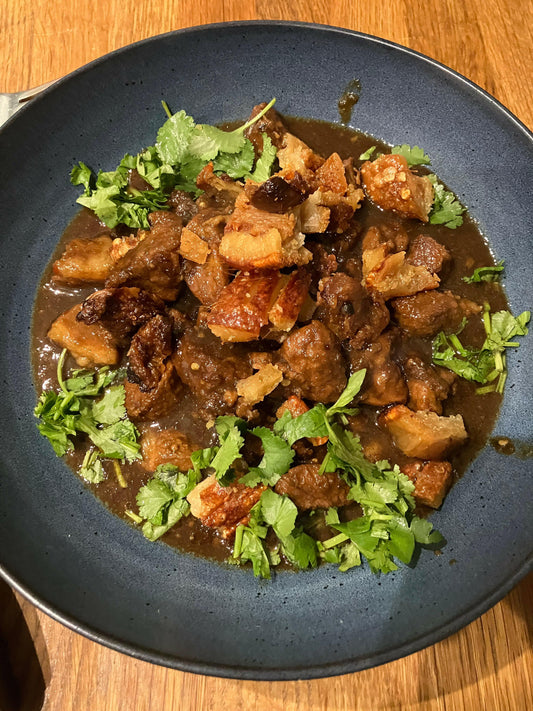 A dark blue bowl of rich meat stew, topped with crispy pork and cilantro, highlights the bold flavors from South Asia on a Plate by Event, inspired by Thai, Malaysian, and Vietnamese cuisine and served on a wooden table.