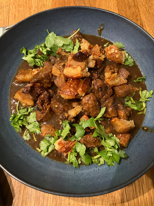 A dark blue bowl of rich meat stew, topped with crispy pork and cilantro, highlights the bold flavors from South Asia on a Plate by Event, inspired by Thai, Malaysian, and Vietnamese cuisine and served on a wooden table.
