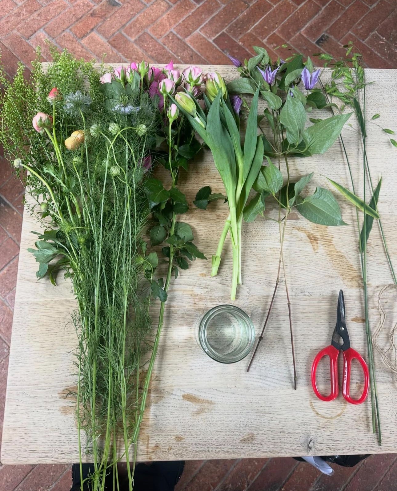 The Art of The Festive Tablescape by Event features a wooden table with flowers, stems, a glass of water, and red-handled scissors—perfect for your next floral workshop. A brick floor forms the backdrop for this creative setup.