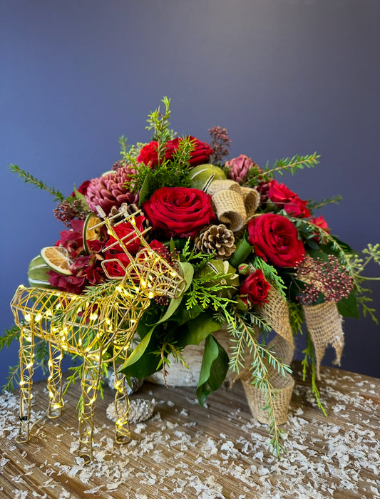 Event’s The Art of The Festive Tablescape features a Christmas floral arrangement with red roses, pinecones, greenery, and burlap ribbon on wood, accented by a gold wire reindeer with lights and imitation snow for a festive touch.