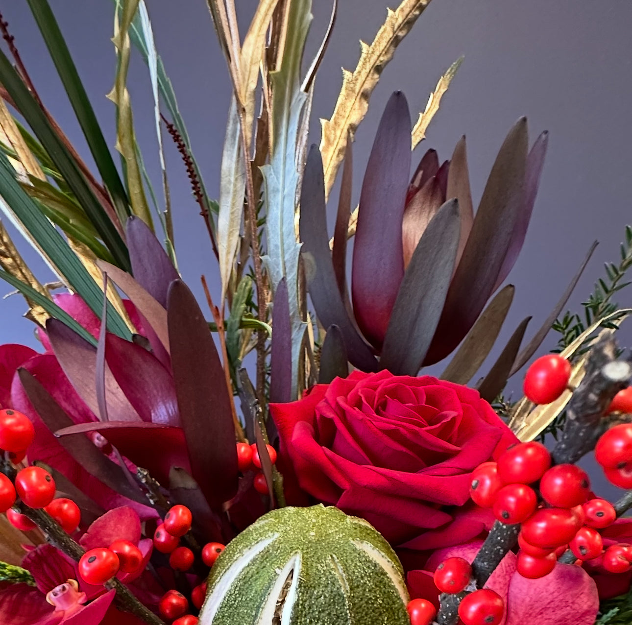 The Art of The Festive Tablescape by Event features a vibrant Christmas floral arrangement with red rose, burgundy protea, red berries, green foliage, and a small green and white ornamental gourd set against a soft gray background.