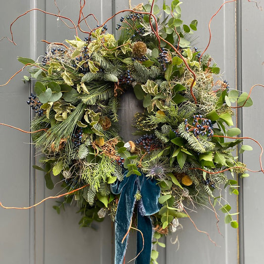 The Event Christmas Wreath Workshop is inspired by a festive wreath of green foliage, pine, berries, twigs, and a dark blue velvet ribbon—perfect for creating your own holiday decoration.