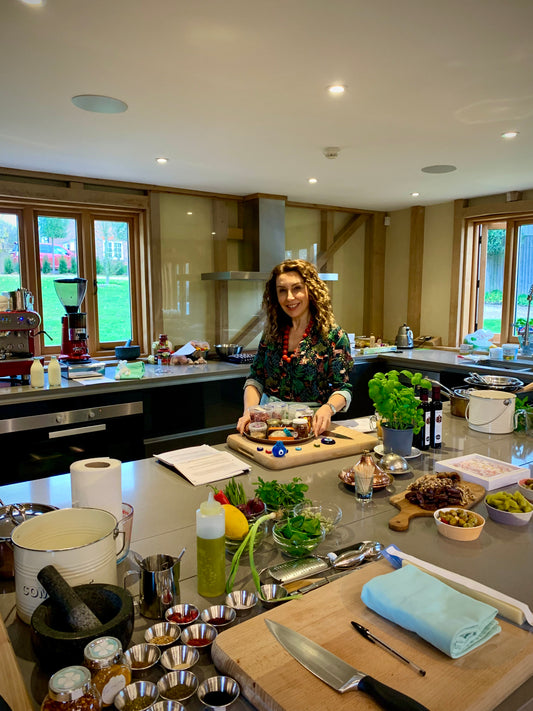 A woman smiles in a modern kitchen, prepping Turkish dishes with fresh herbs on a cutting board using Flavours of the Ottoman Empire by Event. Sunlight streams through large windows, unveiling a lush garden outside.