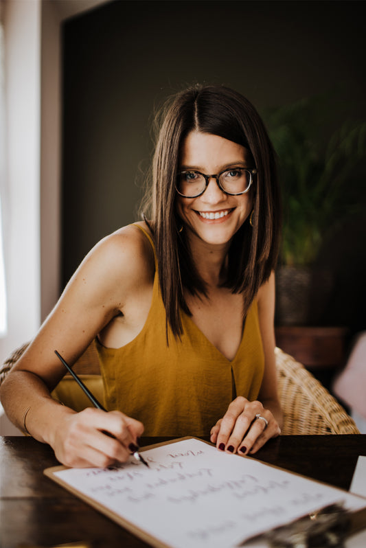 A woman with long brown hair and glasses, in a mustard yellow sleeveless top, smiles while writing on a clipboard at a wooden table—perhaps planning her next beginner project with Modern Calligraphy by Event. A potted plant sits in the background.