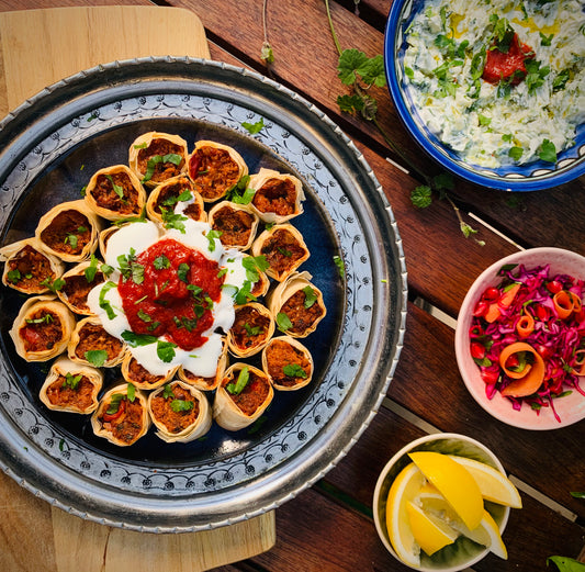 Experience bold vegetarian flavours with Event’s Mastering Bold Vegetarian Flavours—a tray of rolled pastries topped with yogurt and red sauce, served with tzatziki, red cabbage salad, and lemon wedges for an authentic Turkish feast.