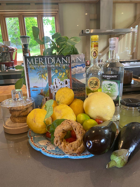 A vibrant display of lemons, tomatoes, eggplants, and pastry from Events Amalfi Coast evokes Italian flavors, set on a kitchen counter with limoncello bottles, honey jar, cookies, and a Meridiani magazine in the background.