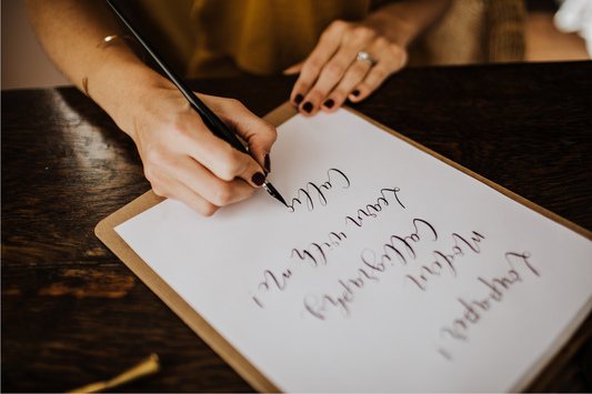 A person with dark nail polish uses the Event Modern Calligraphy set to write elegant cursive letters with a black pen on white paper at a dark wooden table.