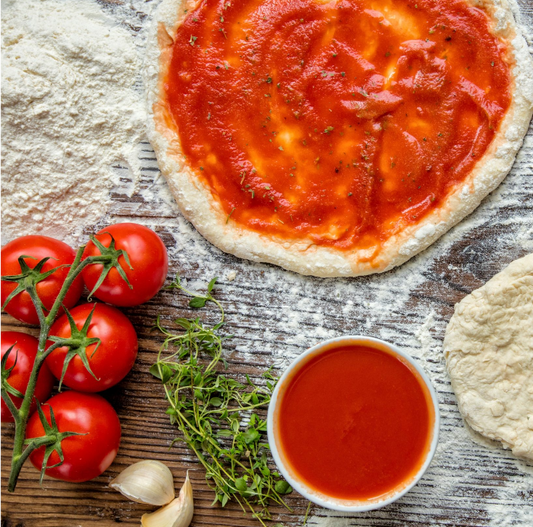 Events Childrens Pizza Making Course features a pizza base with tomato sauce on a floured wooden surface, surrounded by fresh ingredients—ideal for fun kids’ cooking sessions or your next family workshop.
