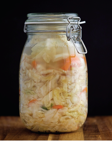 Event’s Fermented Foods Workshop features a glass jar of homemade shredded cabbage and carrot slices, sealed with a metal clasp lid. Perfect for gut health, it sits on a wooden surface against a black background.