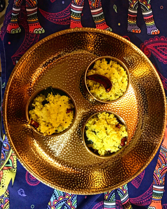 Event’s The Magic of Sri Lankan Cuisine features three small bowls of yellow, shredded coconut dish on a golden hammered metal plate, set atop a colorful patterned cloth with elephant designs.