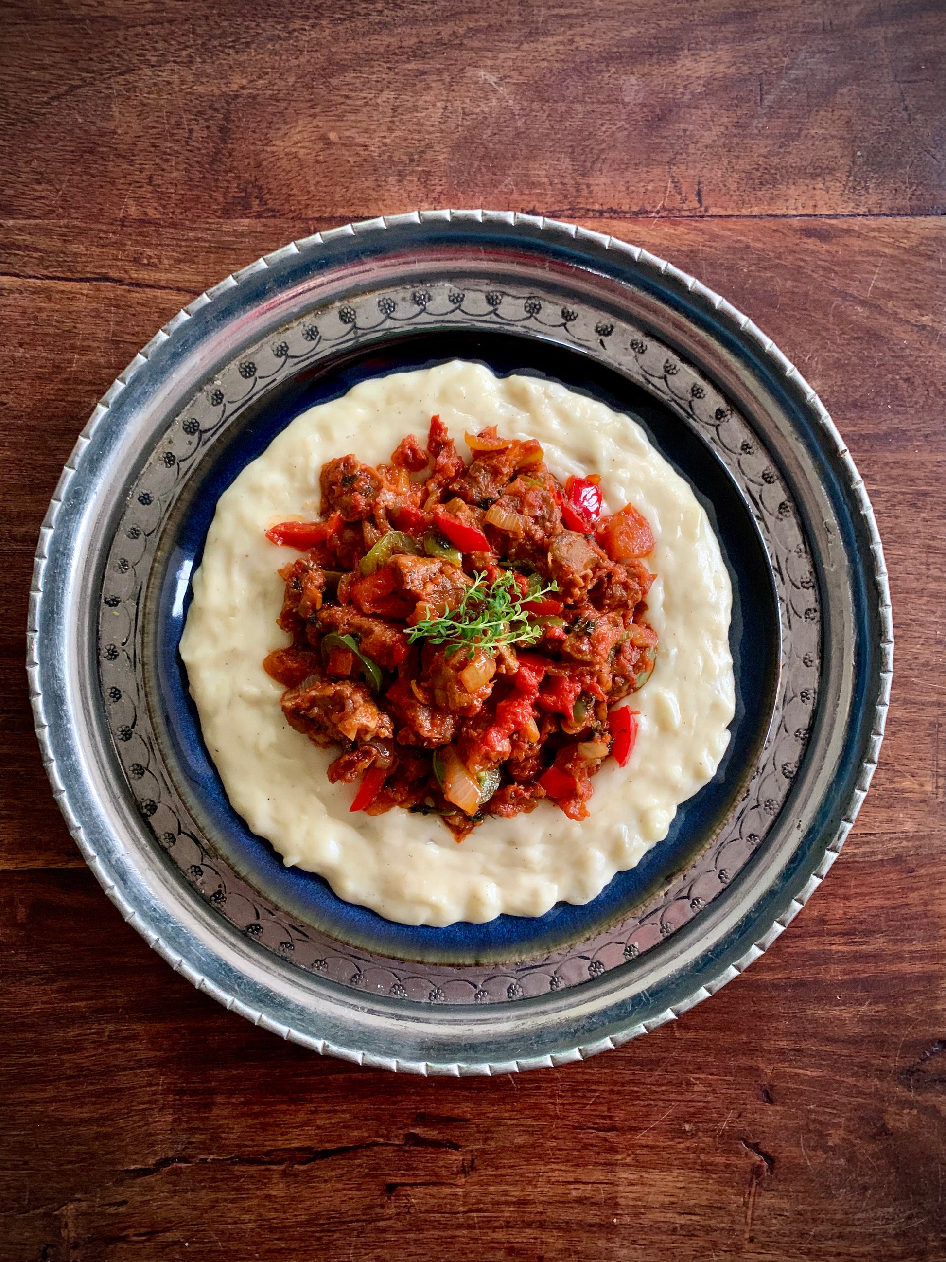Events Flavours of the Ottoman Empire features creamy mashed potatoes topped with chunky tomato and pepper stew, garnished with fresh herbs and served on a decorative metal plate over a wooden table.