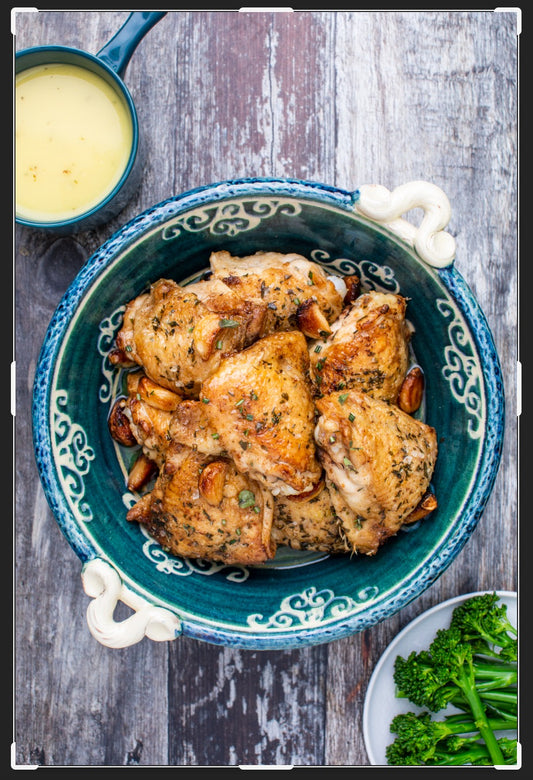 The Event French Bistro features roasted chicken thighs and garlic in a blue patterned bowl, paired with light yellow sauce and steamed broccolini, served on a rustic wooden table for an authentic French dining experience.