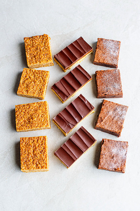 Three rows of Honesty's Afternoon Delight bars on a light surface: golden oat flapjacks left, chocolate-topped Millionaire’s Shortbread center, and powdered sugar bars right. Each row has four pieces.