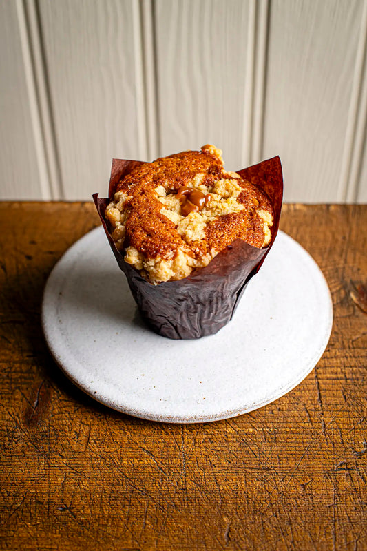 An Honesty Apple & Caramel Muffin in a brown paper liner rests on a white plate over a rustic wooden table, with a light-textured wall in the background.