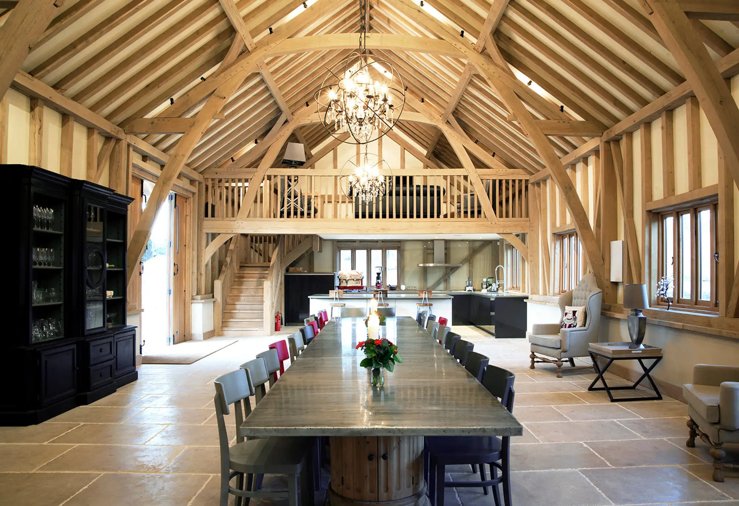 A spacious, rustic dining room with exposed wooden beams, a long dining table surrounded by chairs, two chandeliers, a kitchen at the back, and a mezzanine level above with wooden railings.