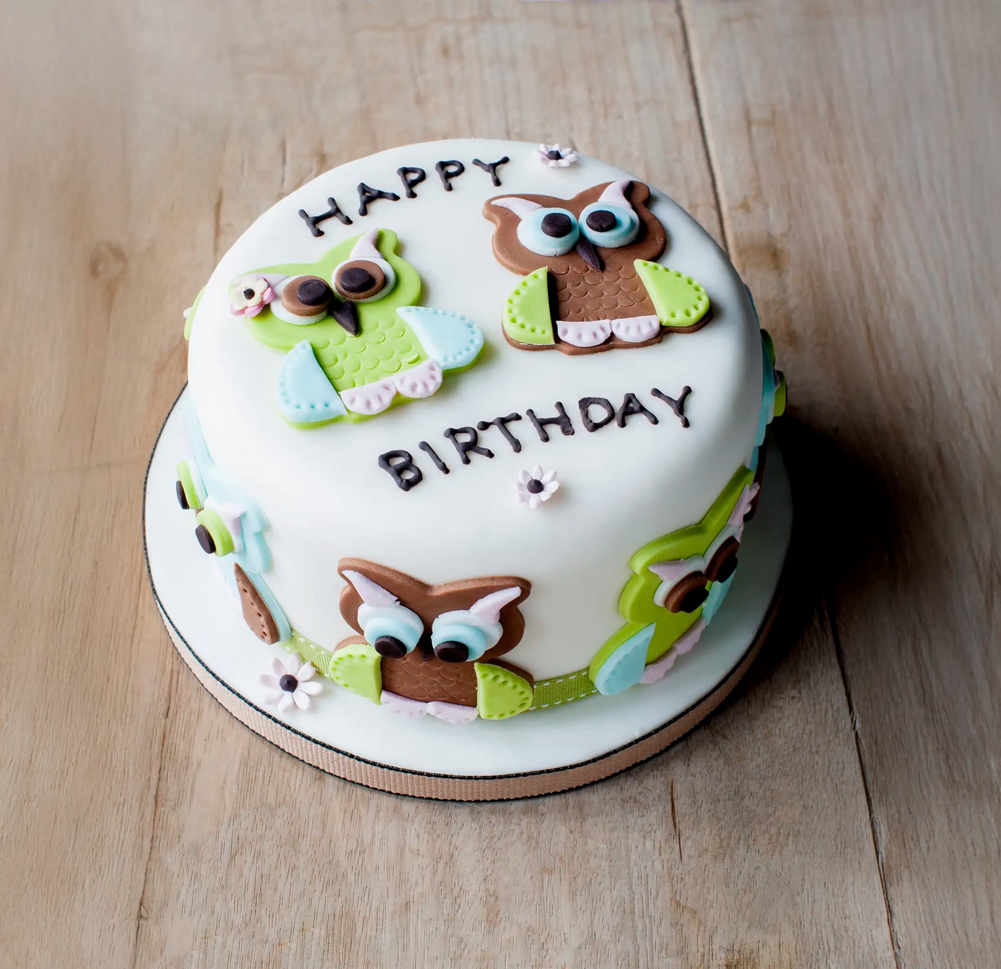 Honesty’s Celebration Cake is a round white birthday cake topped with colorful fondant owls, flowers, and “HAPPY BIRTHDAY”—an elegant centerpiece for any celebration, beautifully displayed on a light wooden surface.