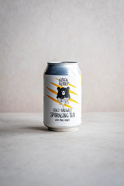 A can of Honesty Cold Brewed Sparkling Tea with Pure Honey rests on a light surface against a neutral background, its label displaying an illustrated bear and bold yellow claw marks.